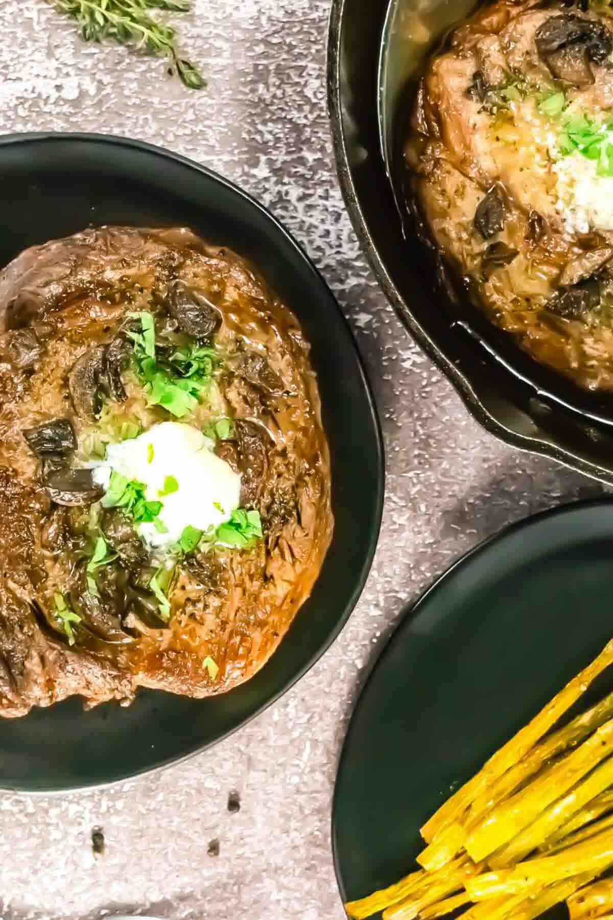 A cooked steak topped with herbs and cream is served on a black plate, with another steak in a pan and a side of asparagus on a separate plate.