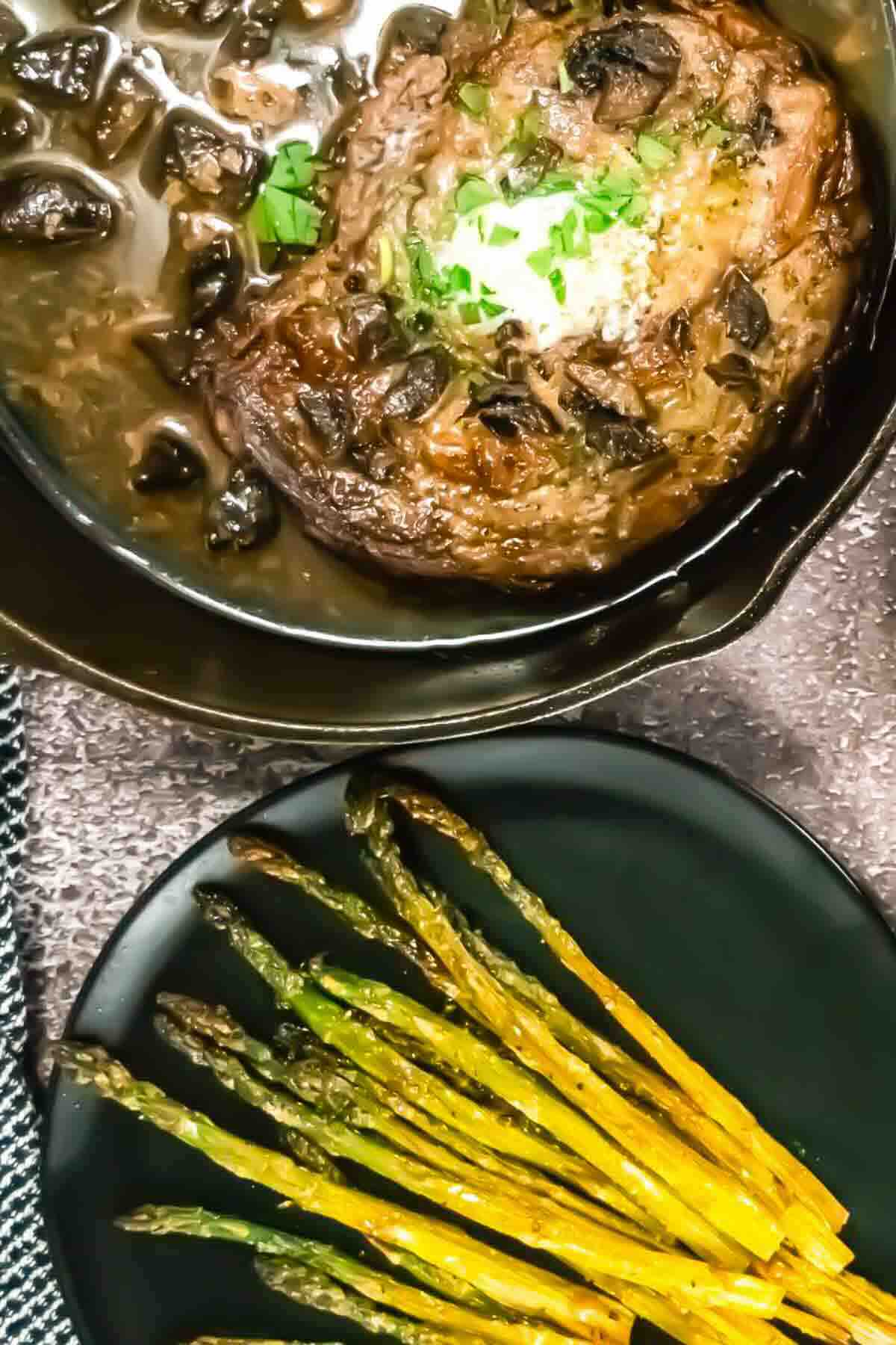 Cooked steak with mushrooms and herbs in a pan, served beside a plate of roasted asparagus spears on a dark surface.
