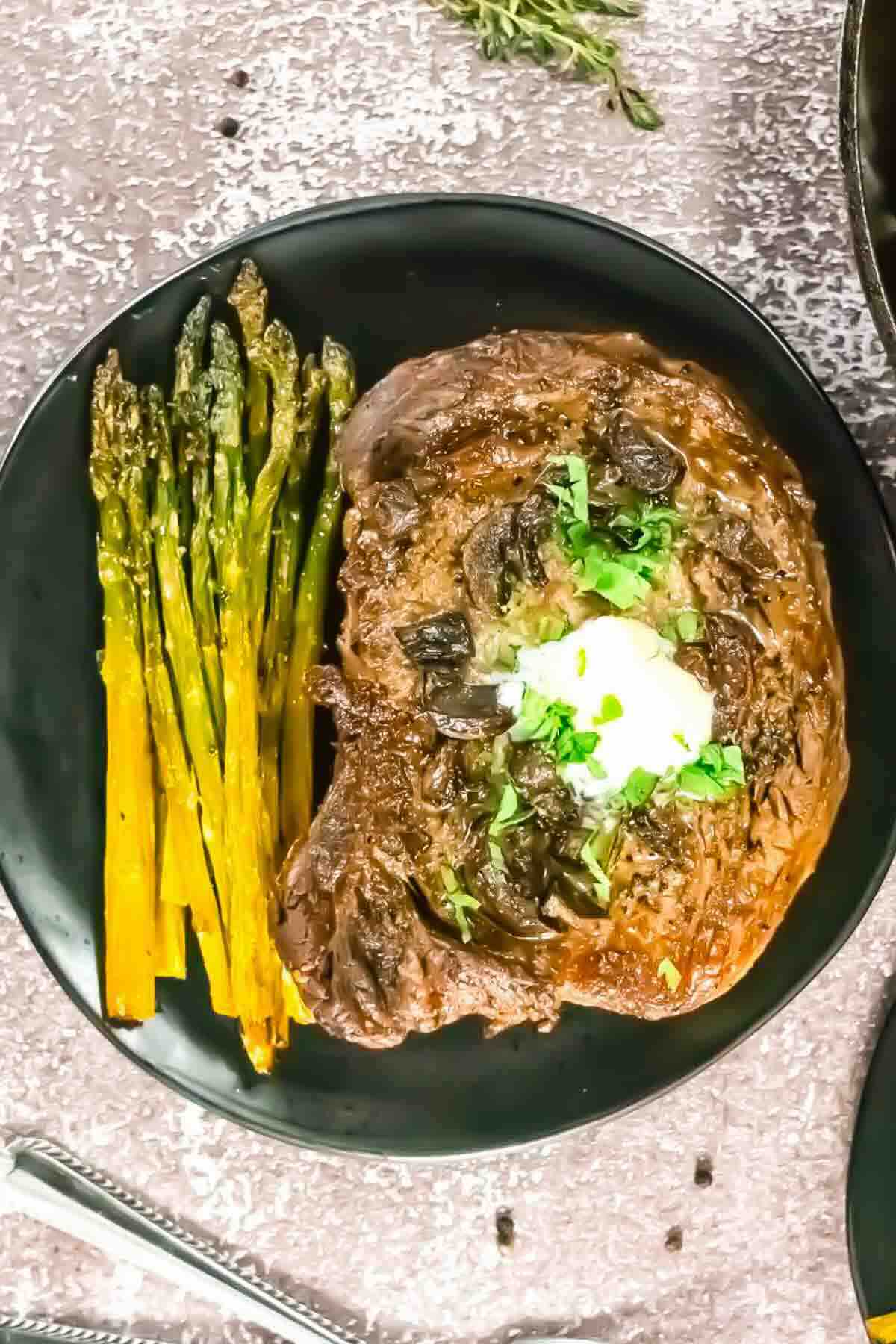 A cooked steak topped with sautéed mushrooms and herbs sits on a black plate next to a serving of roasted asparagus.