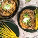 Cooked steak with herbs and butter served on a black plate with roasted asparagus; another steak and more asparagus are nearby on the table.