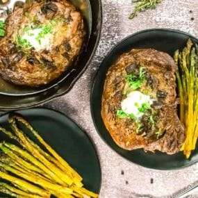 Cooked steak with herbs and butter served on a black plate with roasted asparagus; another steak and more asparagus are nearby on the table.