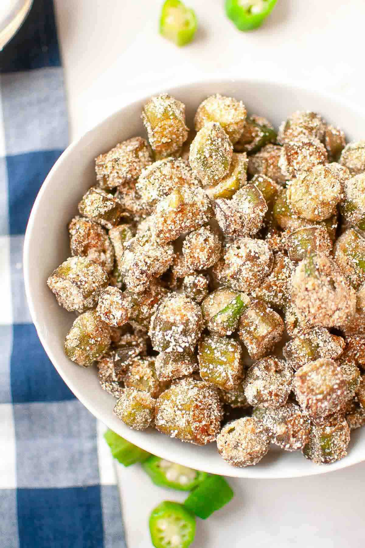 A white bowl filled with breaded and fried okra pieces sits on a blue and white checkered cloth.