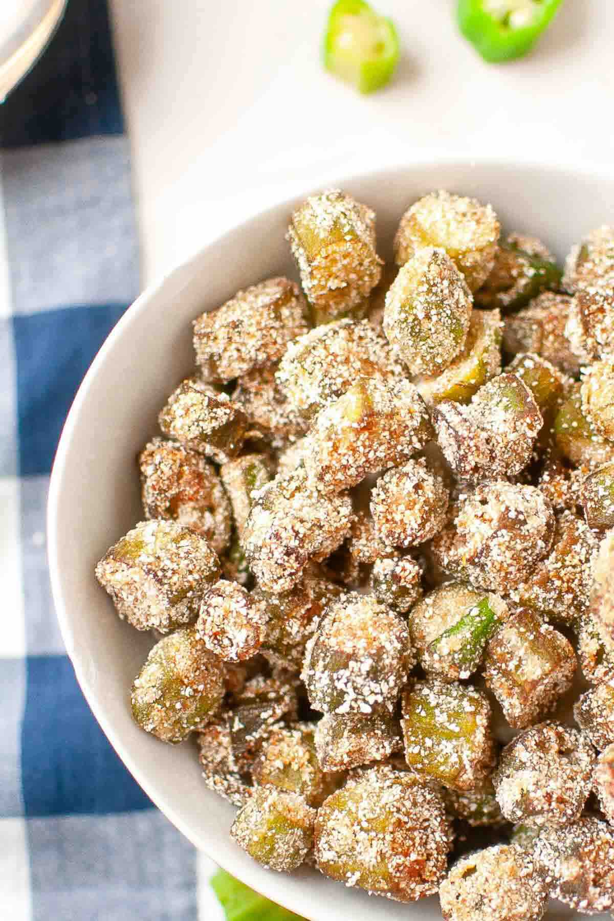 A bowl of fried okra coated in a light layer of seasoning and breadcrumbs, placed on a table with a checkered cloth, with some sliced okra in the background.