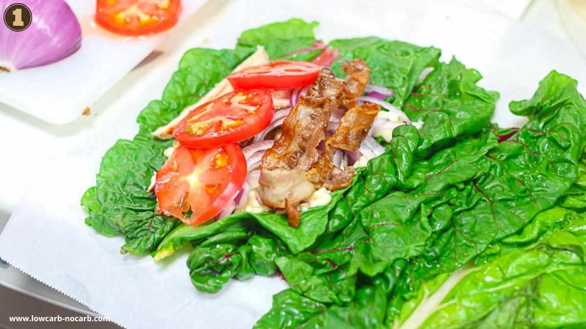 A sandwich wrap with bacon, tomato slices, and onions on large green leafy lettuce, with more greens on the side.