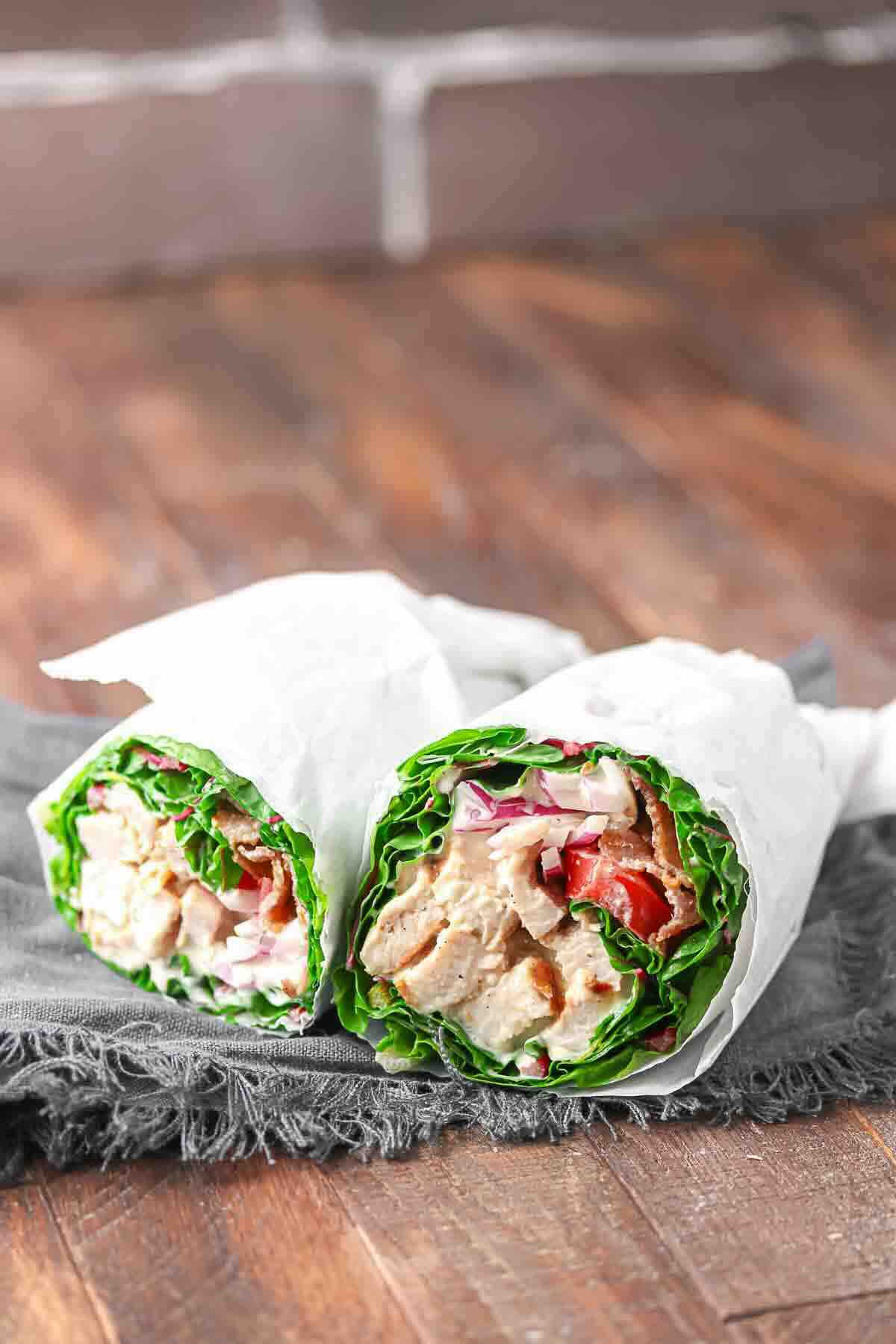 Two lettuce wraps filled with grilled chicken, chopped tomatoes, and sliced red onions, wrapped in white paper, placed on a dark cloth on a wooden surface.