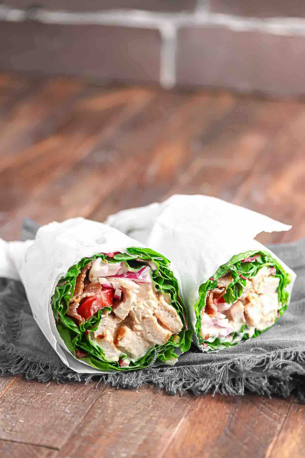 Two halves of a lettuce wrap filled with grilled chicken, sliced tomatoes, onions, and sauce, wrapped in white paper and placed on a dark cloth.