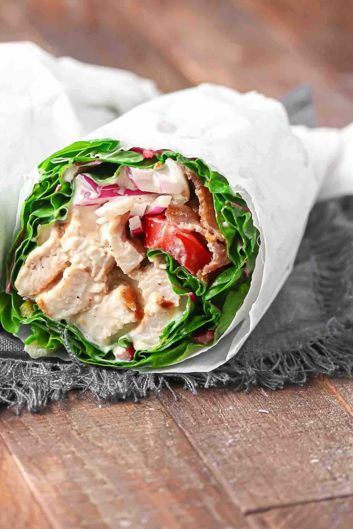 A lettuce wrap filled with sliced grilled chicken, tomatoes, red onions, and sauce, wrapped in white paper on a rustic wooden surface.