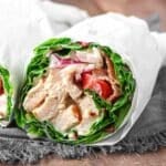 Close-up of a chicken wrap with lettuce, tomatoes, and sauce, wrapped in white paper on a dark cloth surface.