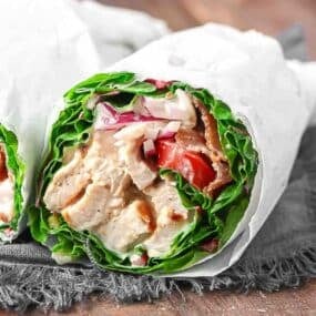 Close-up of a chicken wrap with lettuce, tomatoes, and sauce, wrapped in white paper on a dark cloth surface.