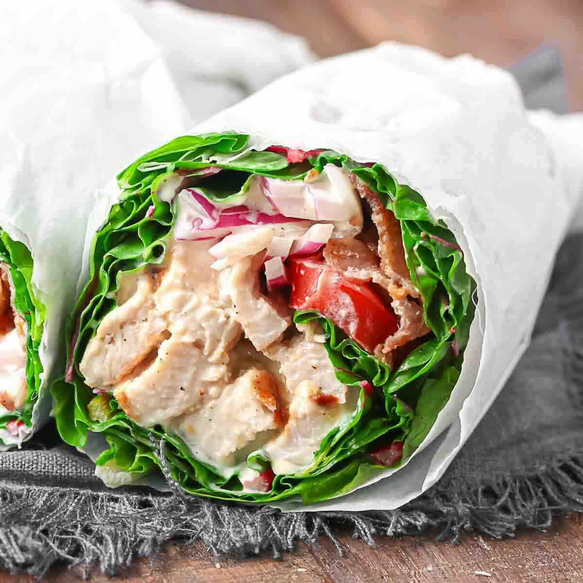 Close-up of a chicken wrap with lettuce, tomatoes, and sauce, wrapped in white paper on a dark cloth surface.