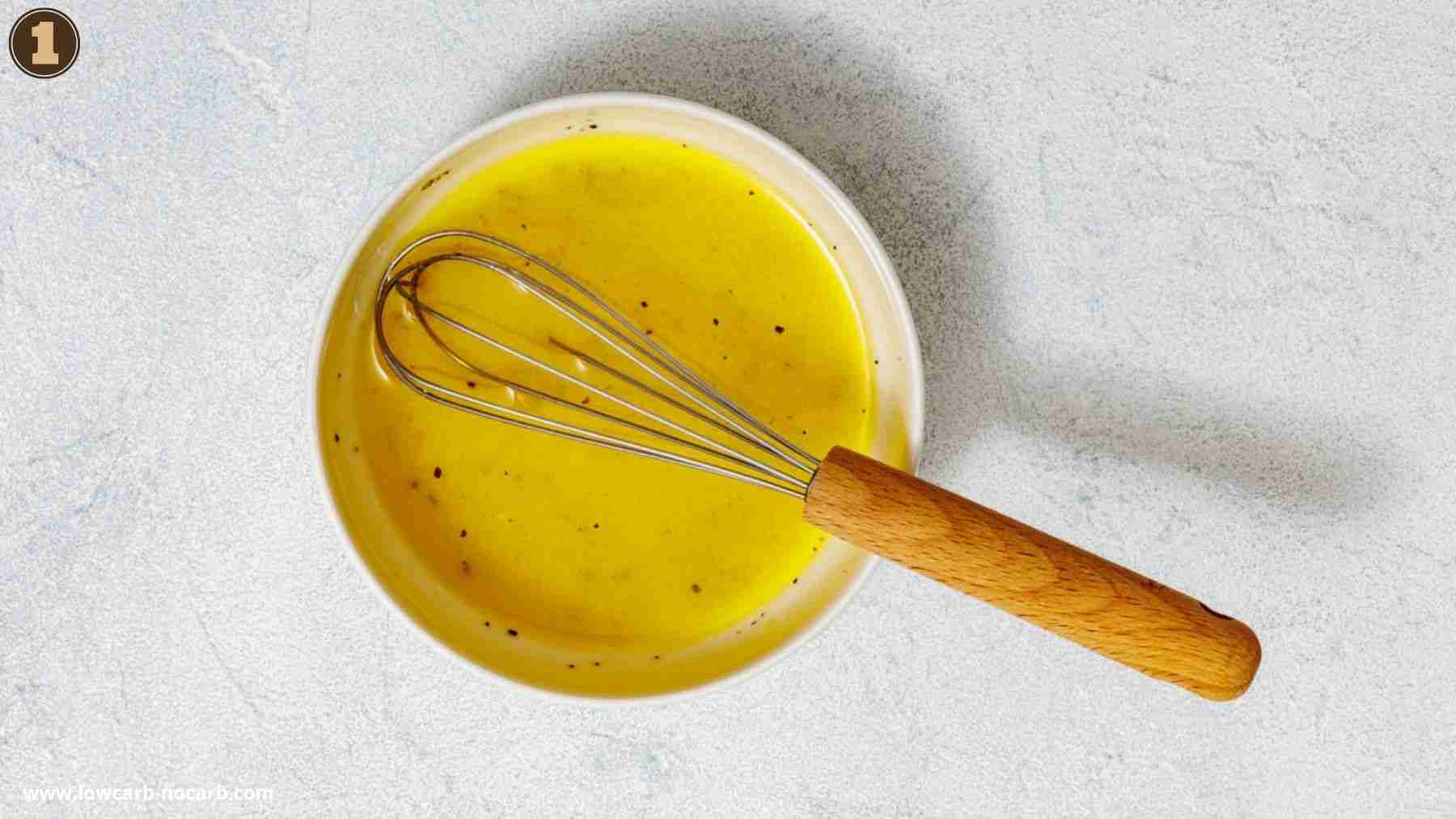 A metal whisk rests in a bowl of yellow liquid with black specks, placed on a light textured surface.
