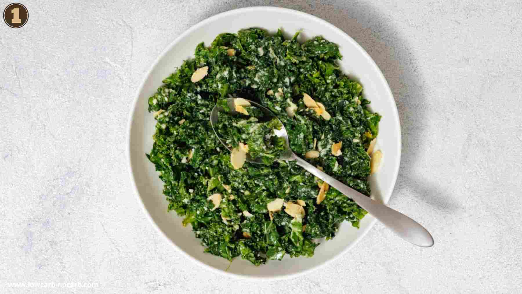A white bowl of chopped kale salad topped with sliced almonds and Parmesan cheese, with a spoon resting inside the bowl, on a light surface.