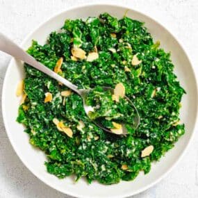 A white bowl filled with chopped kale salad topped with sliced almonds, grated cheese, and a spoon resting inside.