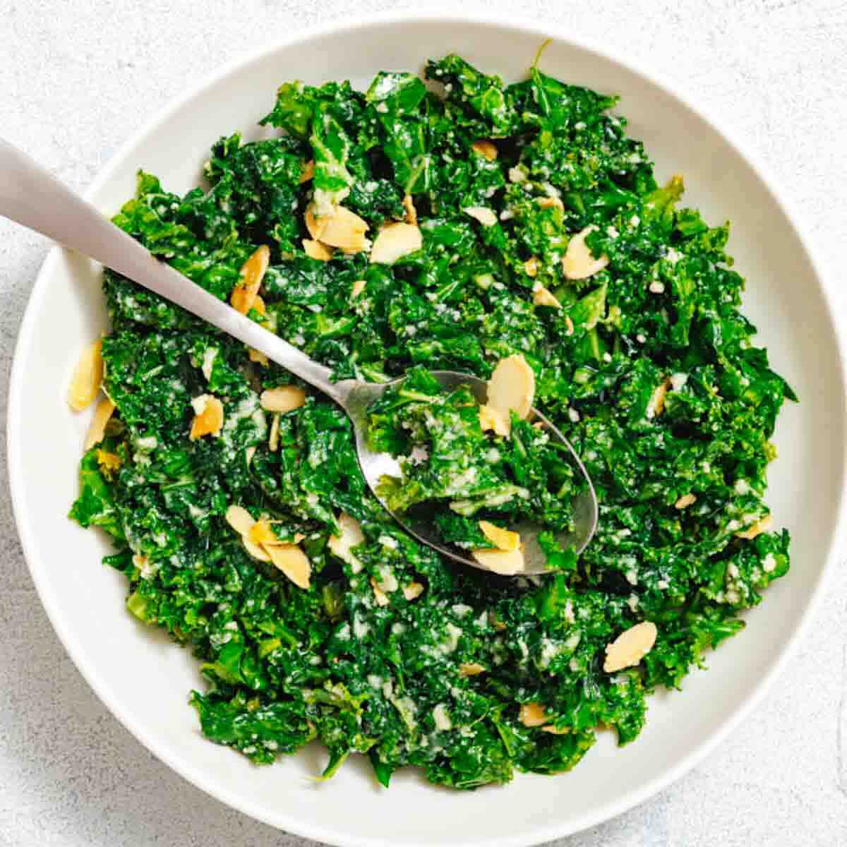 A white bowl filled with chopped kale salad topped with sliced almonds, grated cheese, and a spoon resting inside.