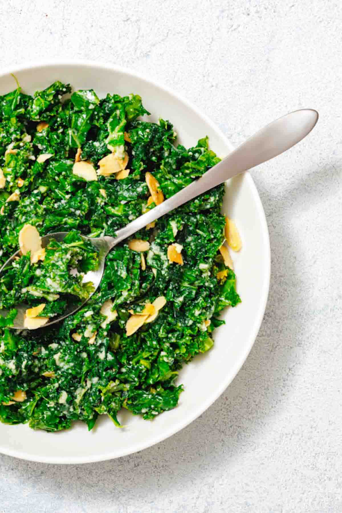 A white bowl filled with kale salad topped with sliced almonds and grated cheese, with a metal spoon resting inside.