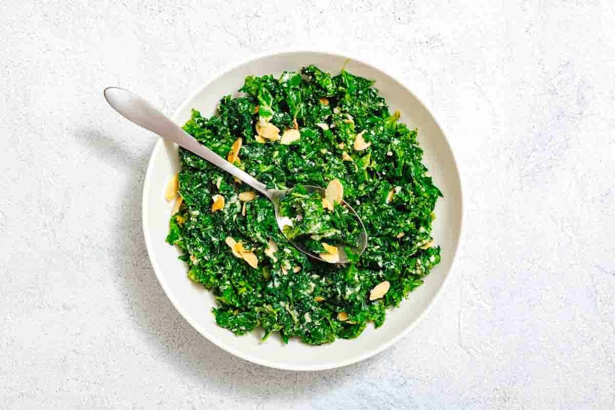 A white bowl filled with chopped kale salad, sprinkled with sliced almonds, and a spoon resting inside, set on a light surface.