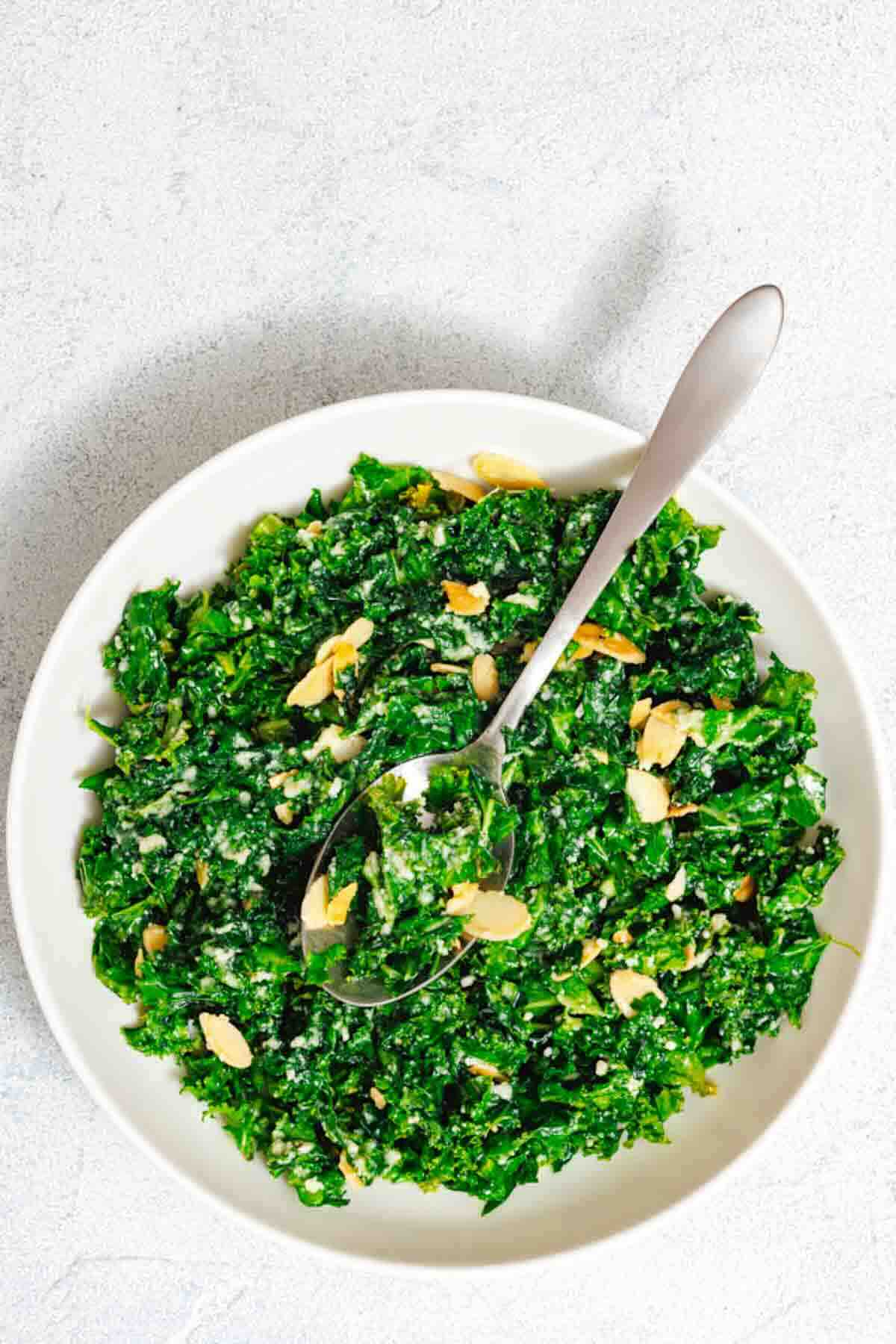 White bowl of chopped kale salad topped with sliced almonds and grated cheese, with a spoon resting inside the bowl.