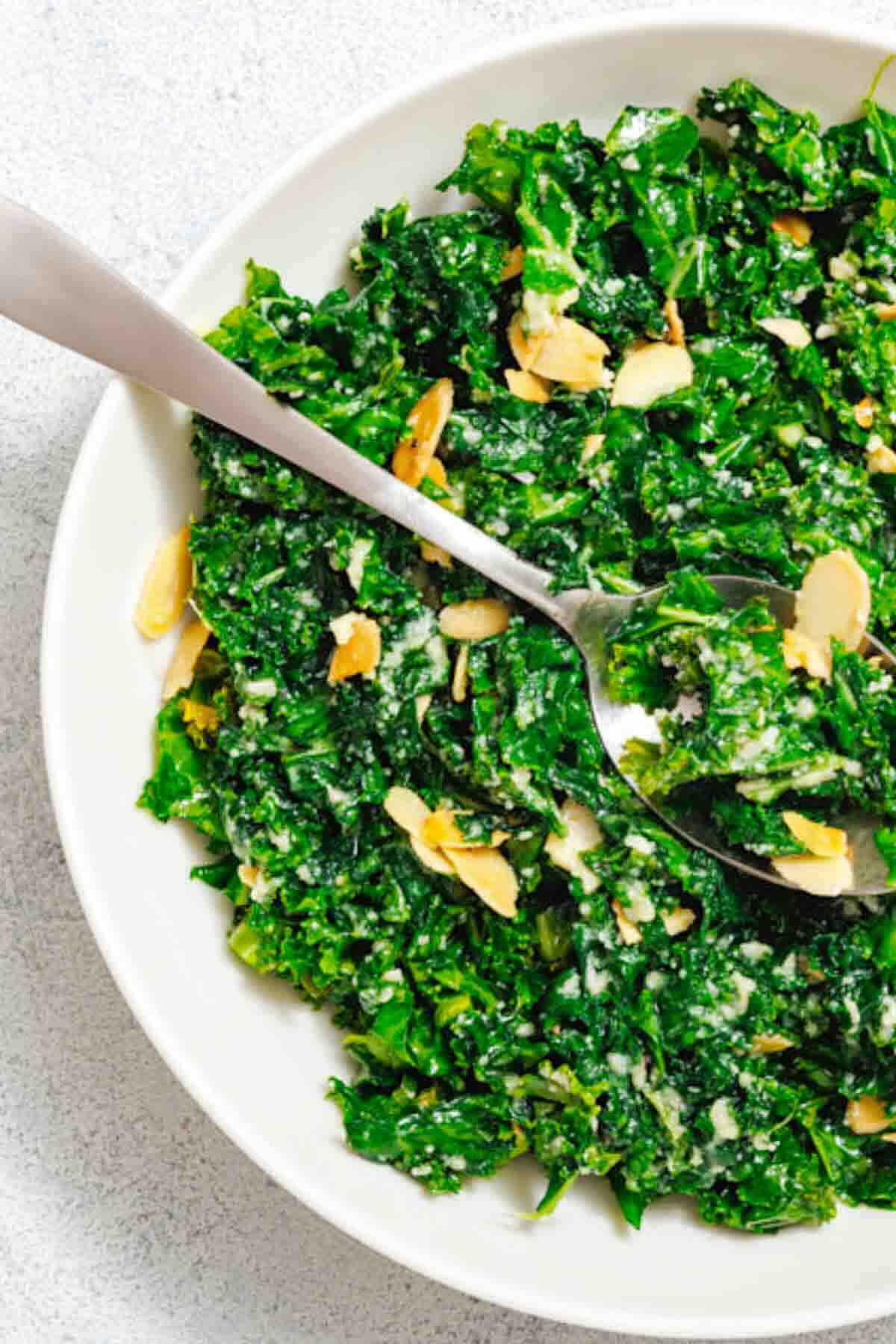 A bowl of kale salad topped with sliced almonds and a creamy dressing, with a serving spoon resting inside.