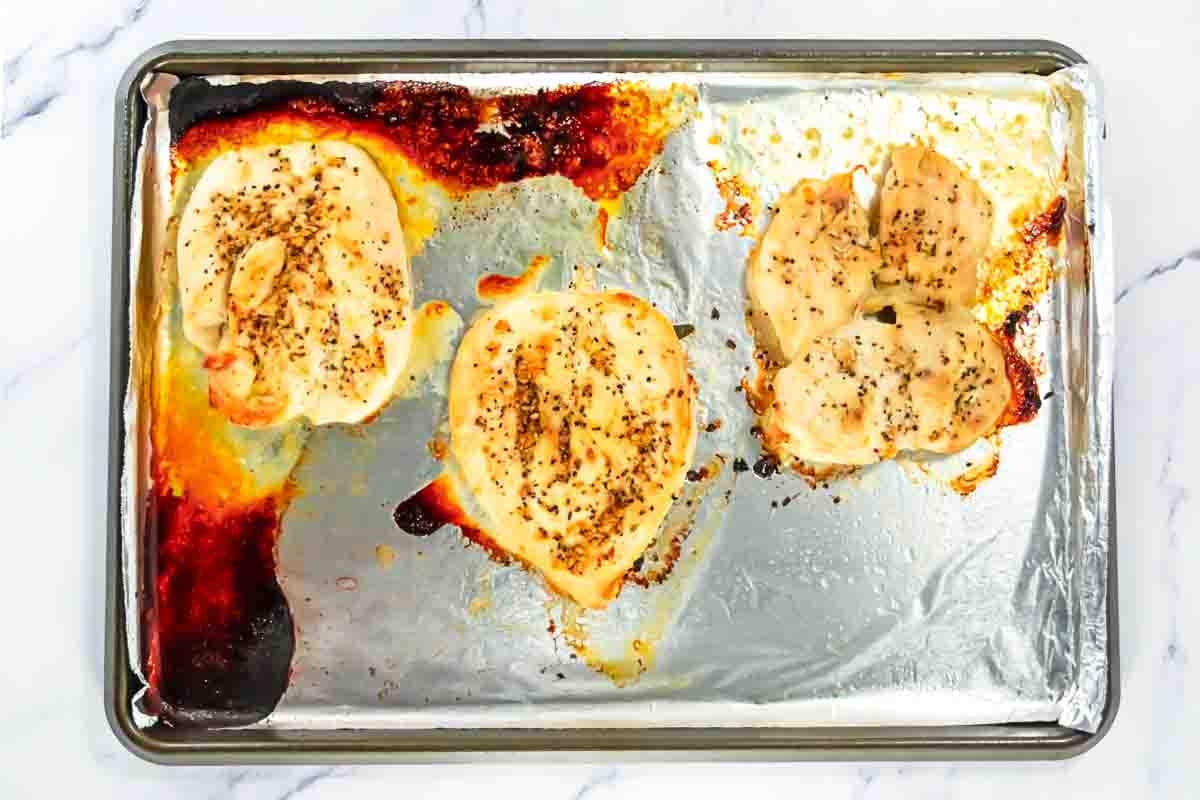 Three seasoned baked chicken breasts on a foil-lined baking sheet with browned edges and some burnt spots on the foil.