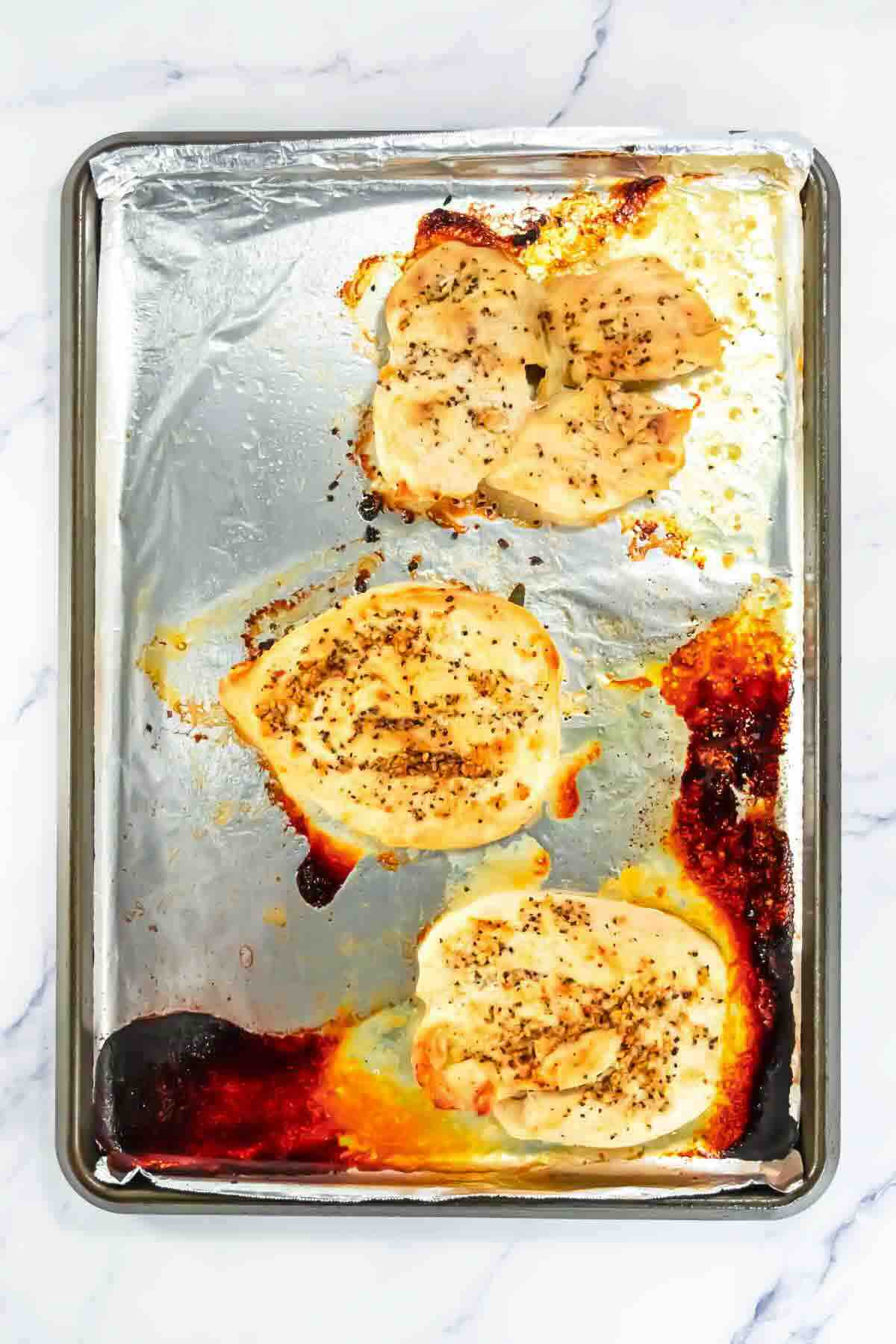 Four baked, seasoned chicken breasts on a foil-lined sheet pan, with browned and caramelized spots on the foil.