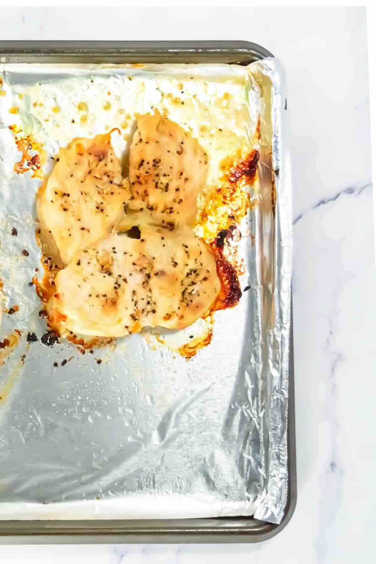 Three baked chicken breasts with seasoning on a foil-lined baking sheet, shown from above on a white marble surface.