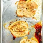 Four baked, seasoned chicken breasts on a foil-lined baking sheet with visible browned spots and juices.