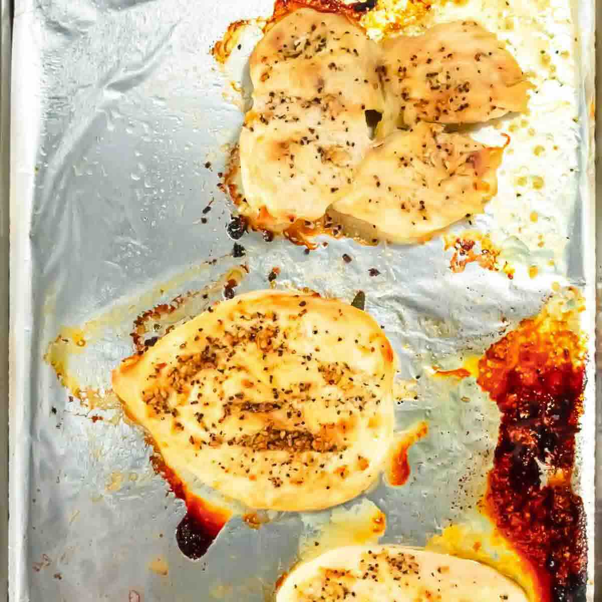 Four baked, seasoned chicken breasts on a foil-lined baking sheet with visible browned spots and juices.