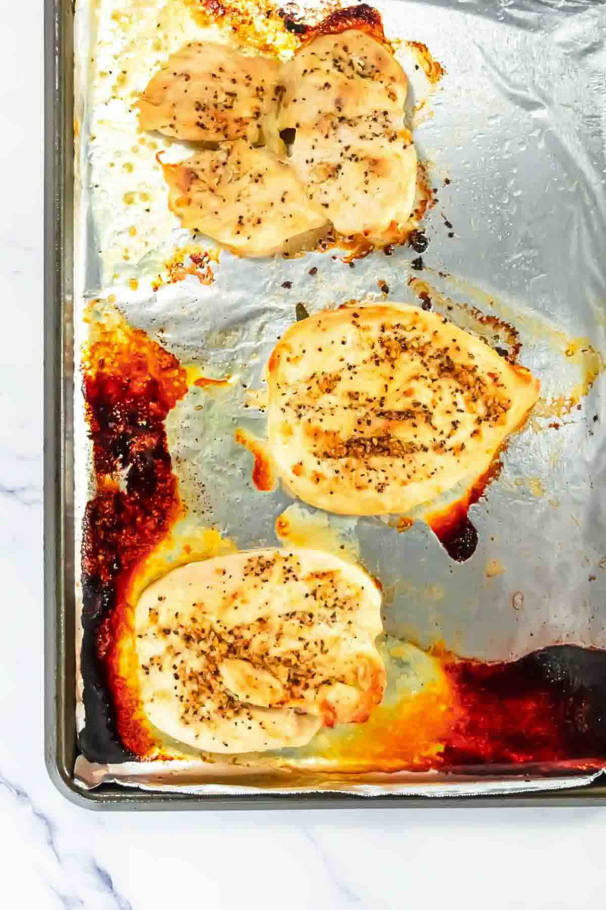 Three pieces of baked, seasoned chicken breast on a foil-lined baking sheet with some browned juices and seasoning visible.