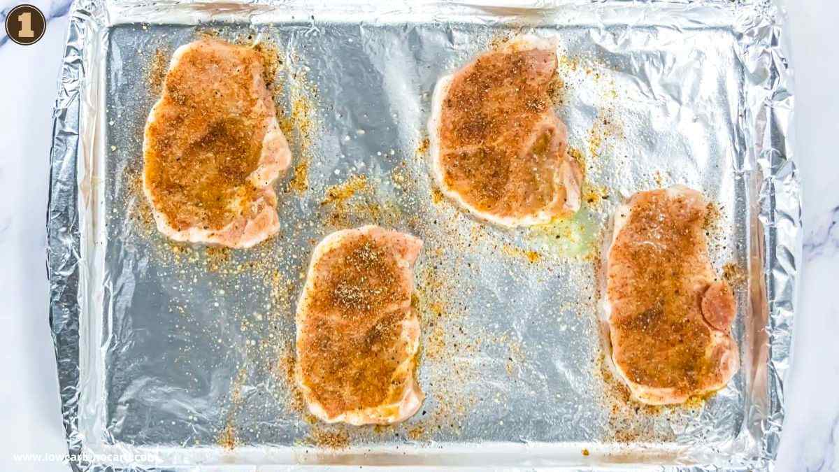 Four raw pork chops coated with seasoning are arranged on a foil-lined baking sheet.