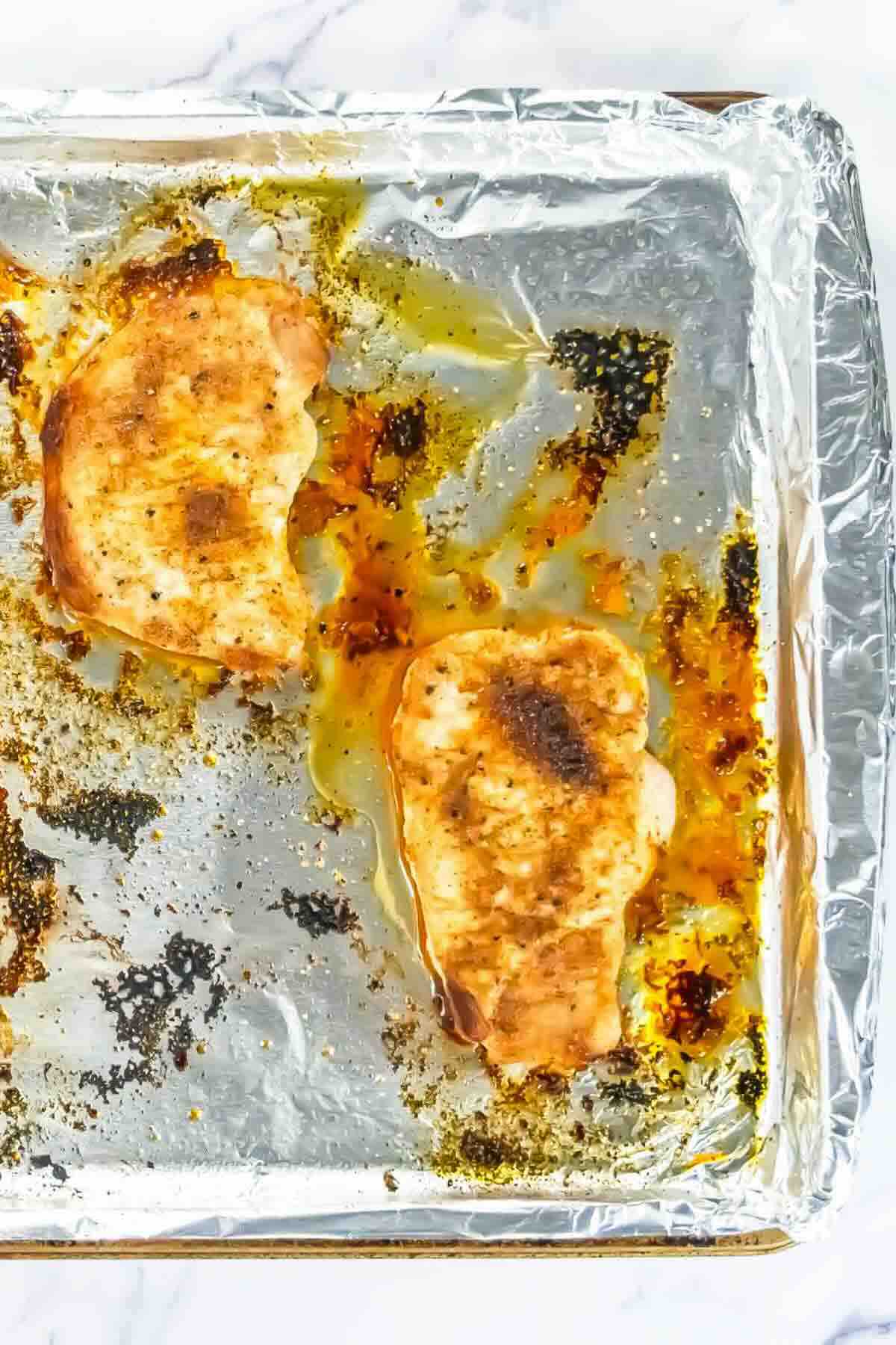 Two cooked, seasoned chicken breasts on a foil-lined baking sheet with oil and browned bits.