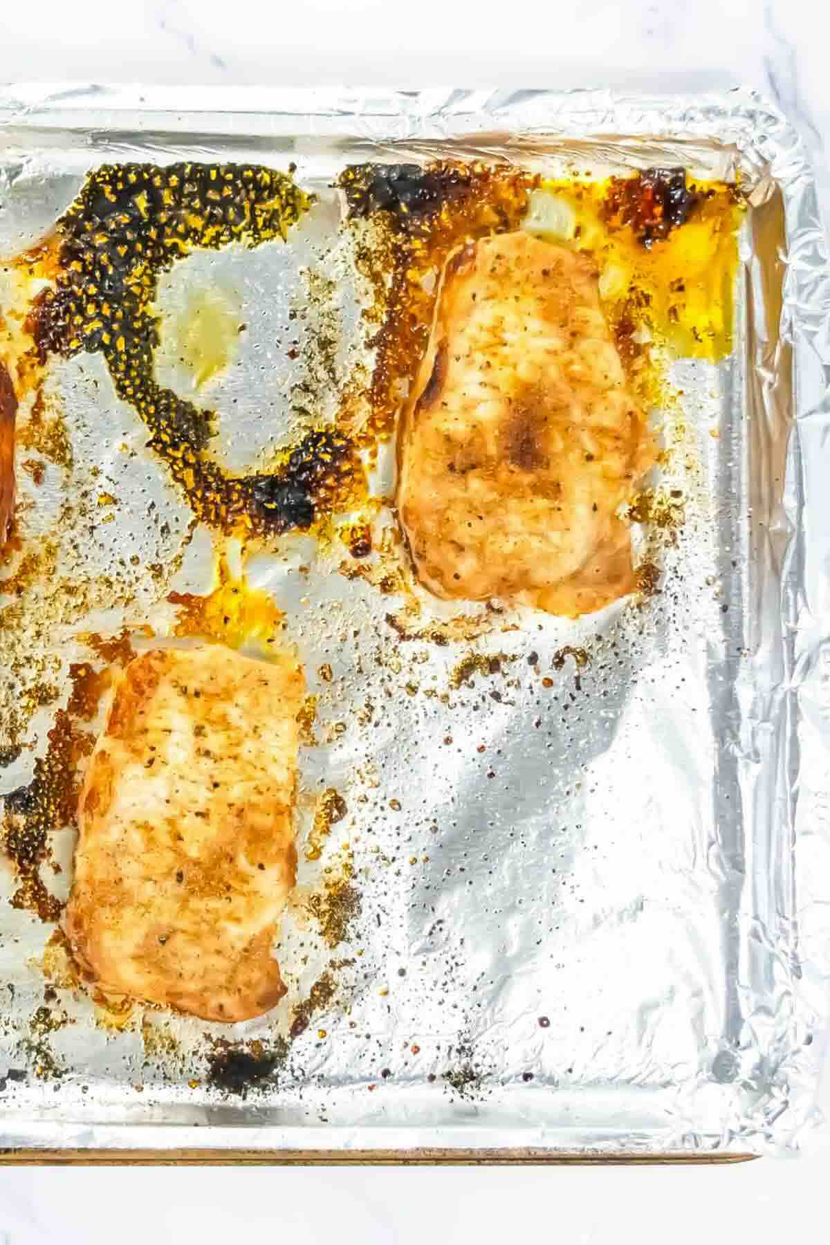 Two cooked, seasoned chicken breasts on a foil-lined baking sheet with some cooking residue visible.