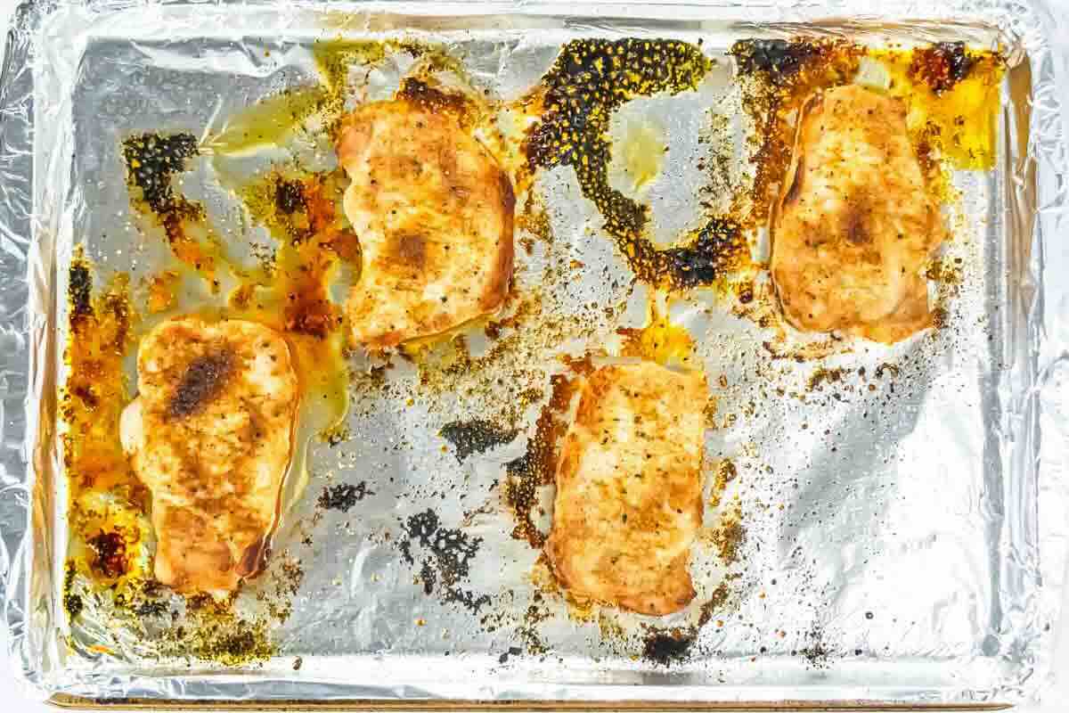 Four cooked chops on a foil-lined baking sheet with browned seasoning and oil residue visible around the meat.