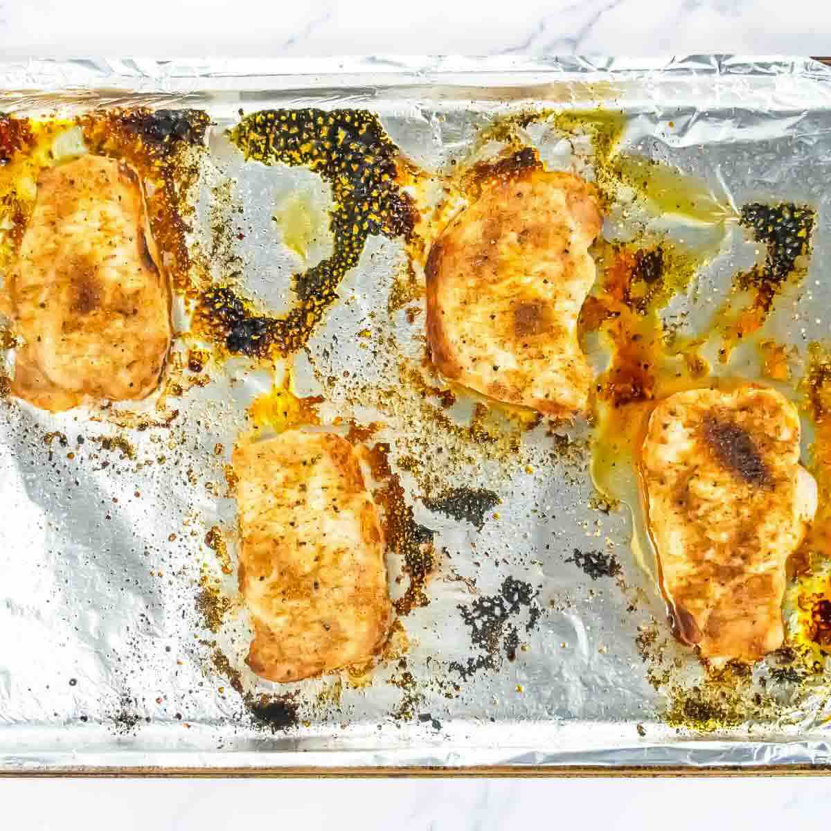 Four cooked pork chops with seasoning on a foil-lined baking sheet, with juices and browned spots visible.