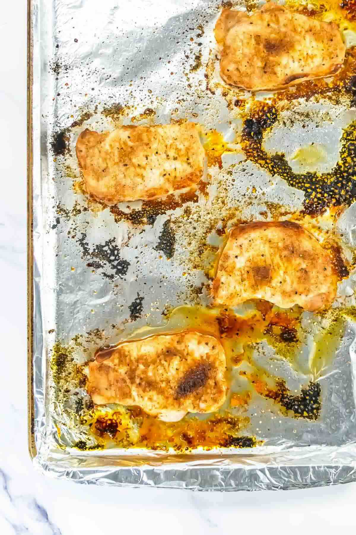 Four seasoned, baked chicken breasts sit on a foil-lined baking sheet with visible oil and seasoning residue.