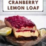 A sliced loaf of cranberry lemon bread with cranberry topping sits on a wooden board, garnished with lemon halves and a cranberry, against a blurred background.