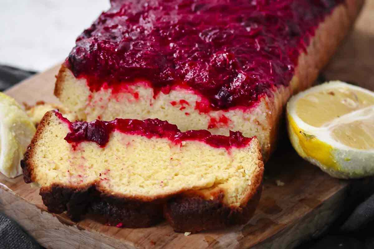 A loaf of cake with cranberry topping is sliced on a wooden board, with two lemon halves placed nearby.