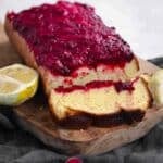 A loaf cake topped with berry compote sits on a wooden board, with two lemon halves placed nearby.
