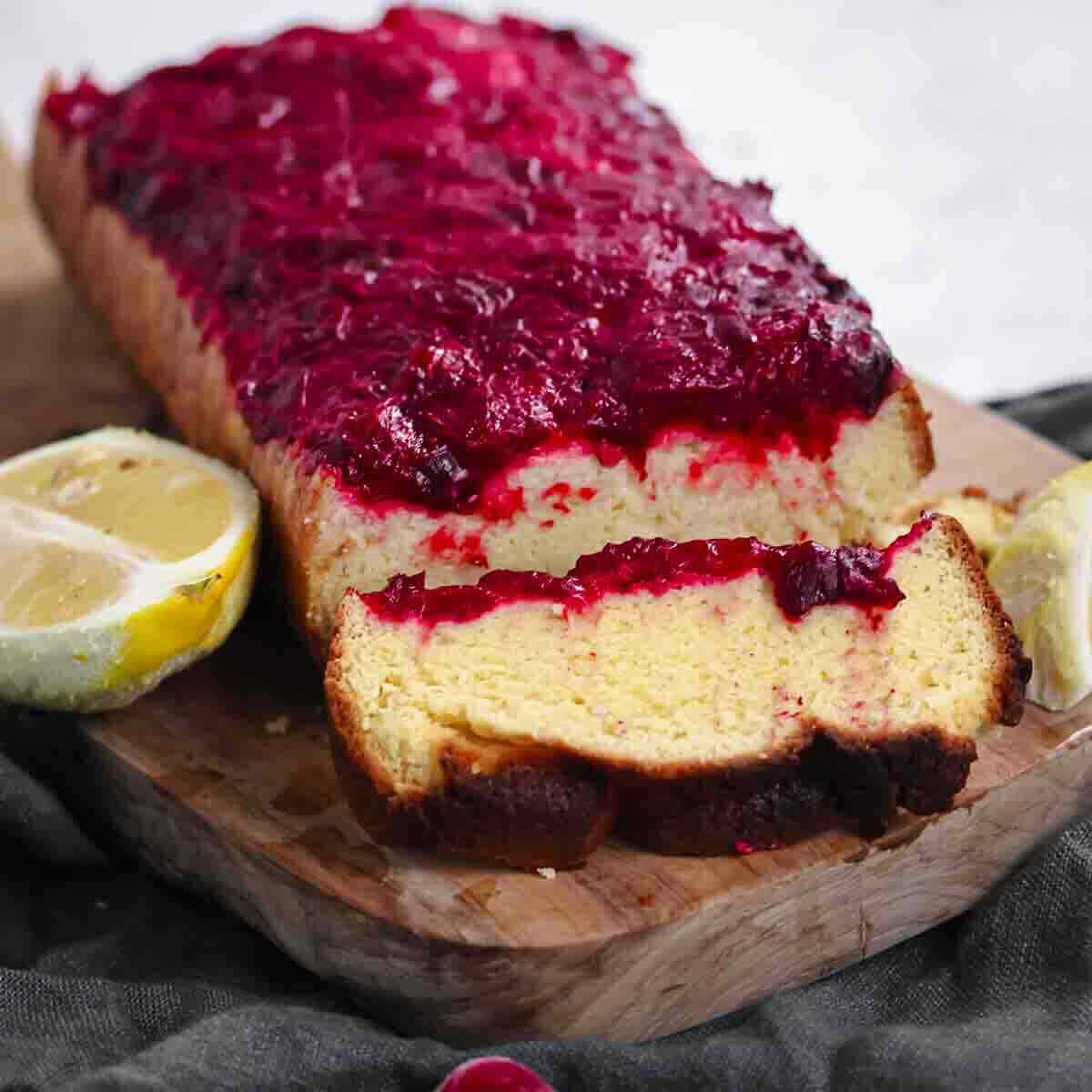 A loaf cake topped with berry compote sits on a wooden board, with two lemon halves placed nearby.