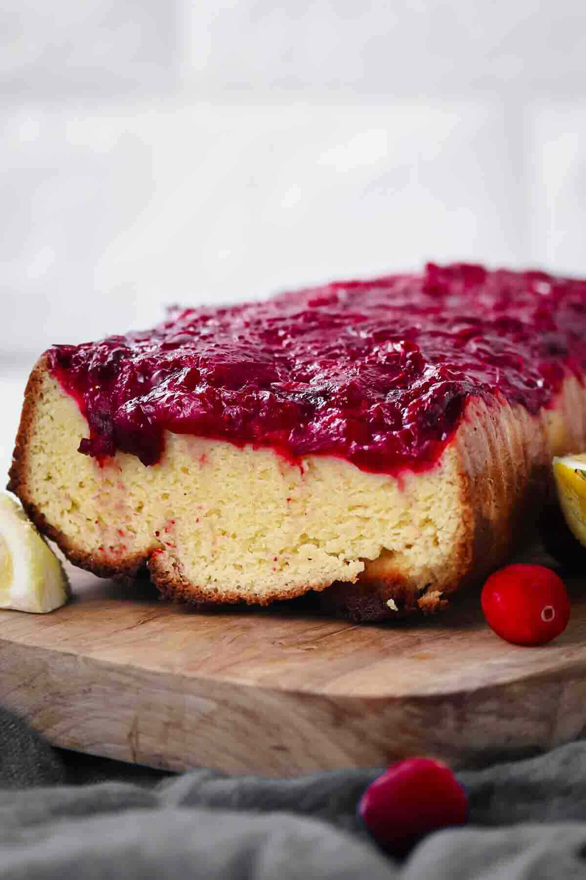 A loaf of cake with a thick layer of cranberry topping sits on a wooden board, with lemon slices and cranberries nearby.