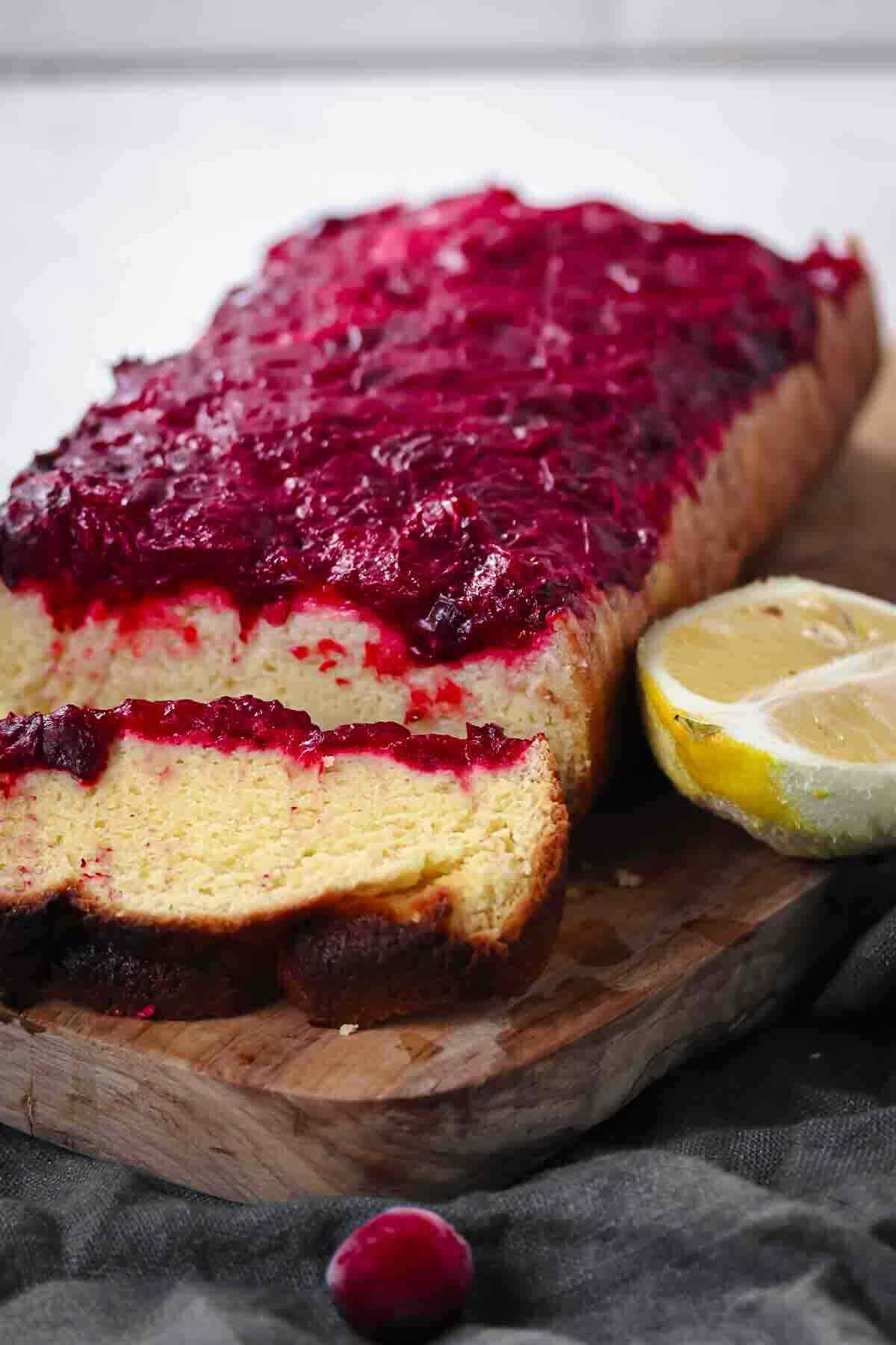 A loaf of cake topped with a layer of red berry sauce, sliced to show the inside, sits on a wooden board next to a halved lemon.