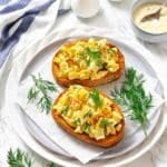 Two slices of toasted bread topped with egg salad and fresh dill are served on a white plate, with extra dill garnish on the side.