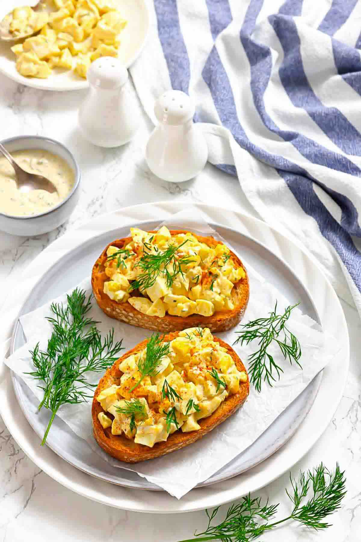 Two slices of toast topped with egg salad and fresh dill are served on a white plate, with a striped cloth, sauce, and salt and pepper shakers nearby.