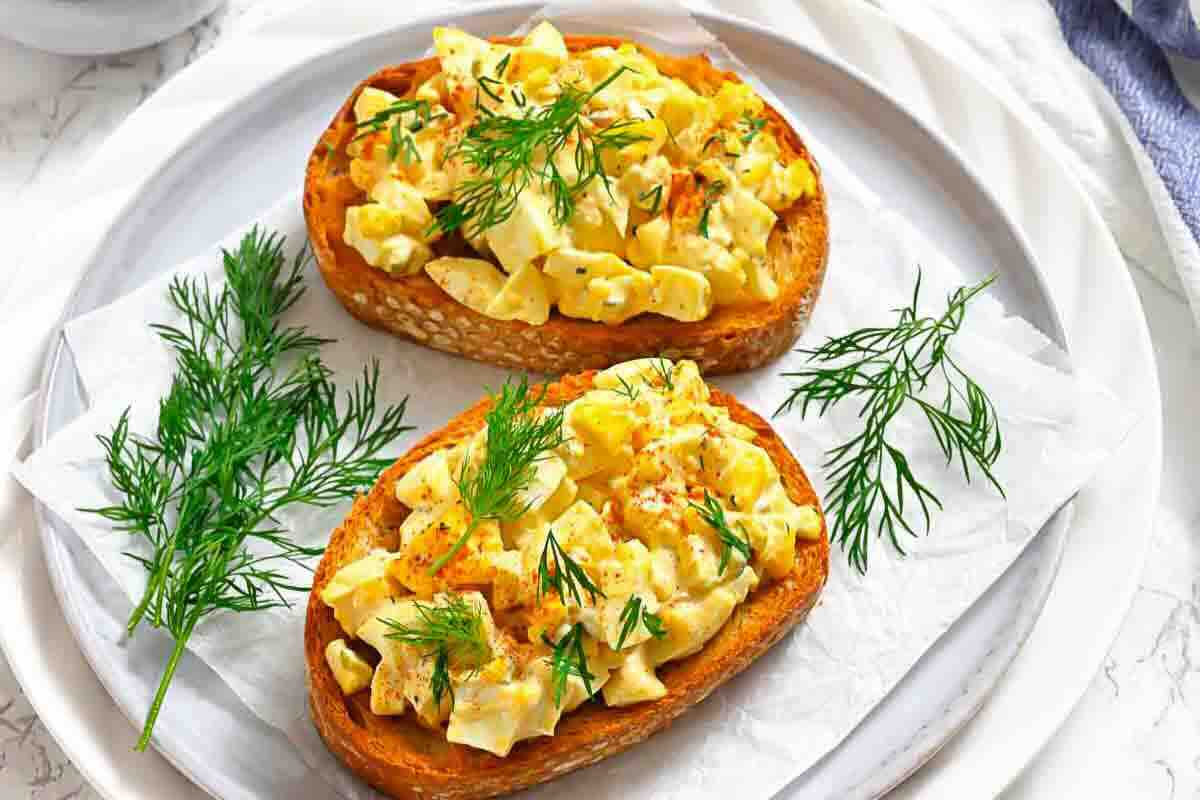 Two slices of bread topped with deviled egg spread and fresh dill are served on a white plate with extra dill garnish.