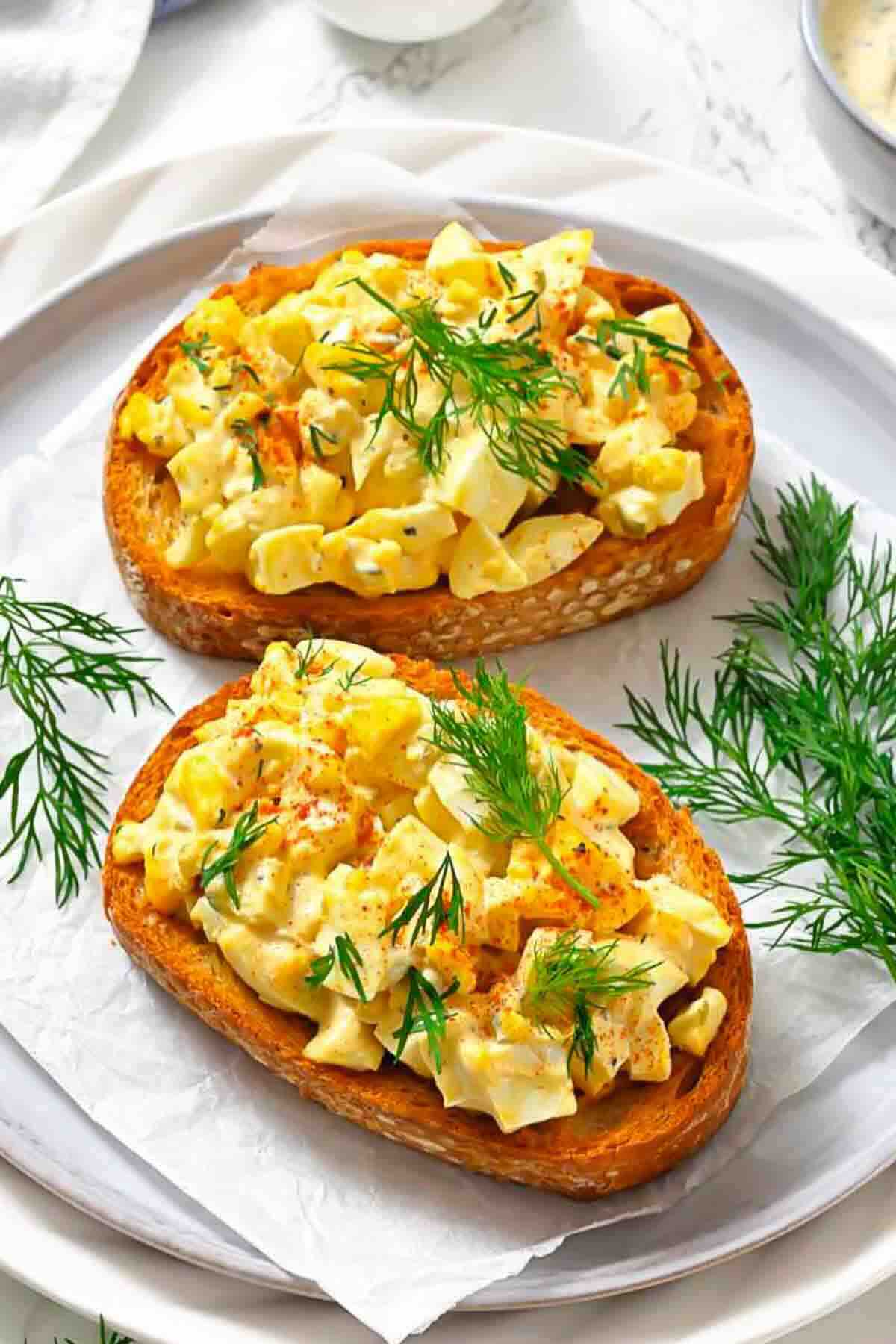 Two slices of toasted bread topped with egg salad and garnished with fresh dill, served on a white plate with extra dill sprigs.
