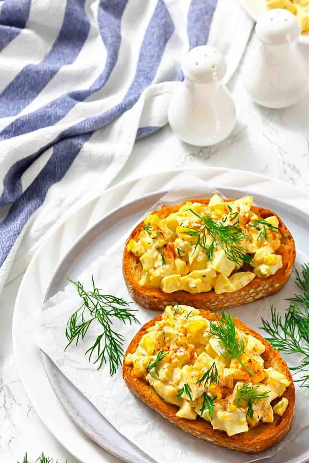 Two slices of bread topped with egg salad and fresh dill, served on a white plate with a sprig of dill, next to a striped cloth and salt and pepper shakers.