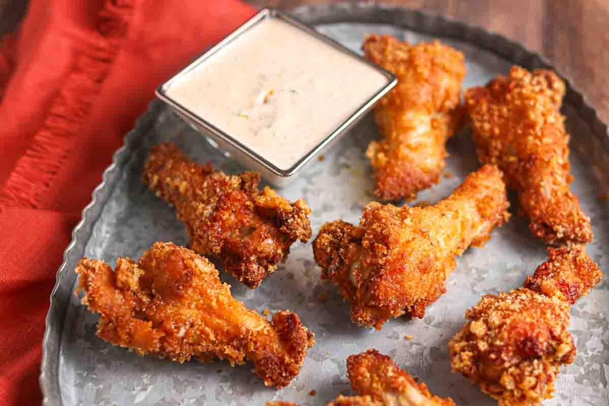 Six crispy fried chicken drumsticks are arranged on a metal tray next to a square dish of dipping sauce, with a red cloth partially visible.