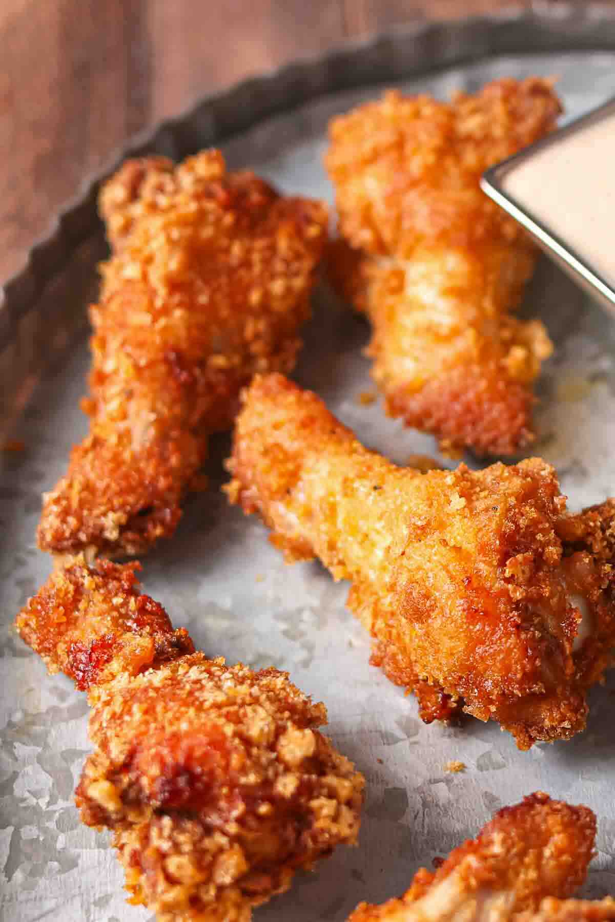 Five pieces of crispy fried chicken wings are arranged on a metal tray next to a small container of dipping sauce.
