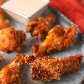 Close-up of several crispy fried chicken wings on a metal tray with a small container of dipping sauce in the background.
