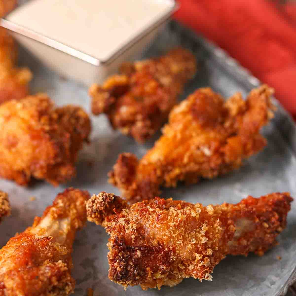 Close-up of several crispy fried chicken wings on a metal tray with a small container of dipping sauce in the background.