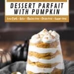 A glass layered with pumpkin dessert and whipped cream, topped with a dusting of spice, next to a gold spoon. Text overlay reads: “Delicious Keto Dessert Parfait with Pumpkin.”